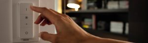 Kitap raflarıyla dekore edilmiş **rahat bir oturma odasında** ortam aydınlatmasını kontrol eden **Philips akıllı LED dimmer anahtarını ayarlayan bir el**.